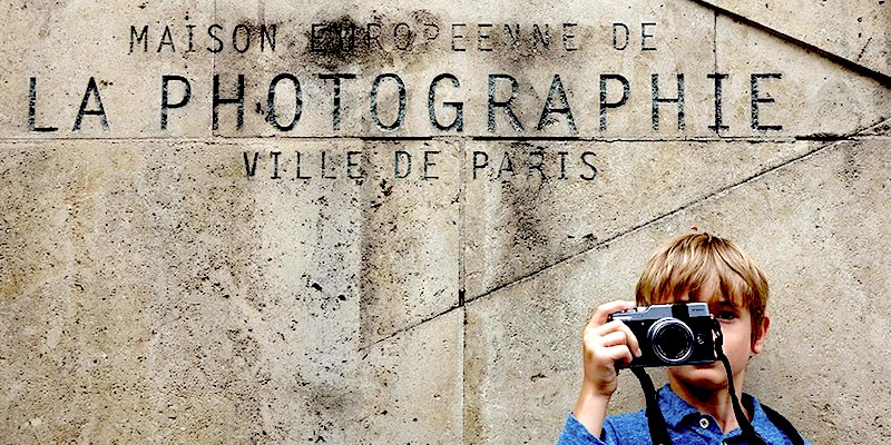 A young boy points his camera at the viewer in from the the sign for La Maison Européenne de la Photographie