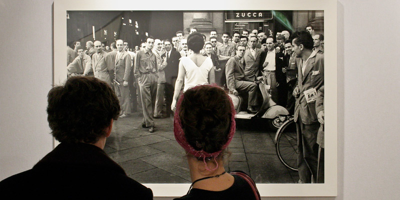 Two visitors look a photograph of men looking at a woman in a white dress