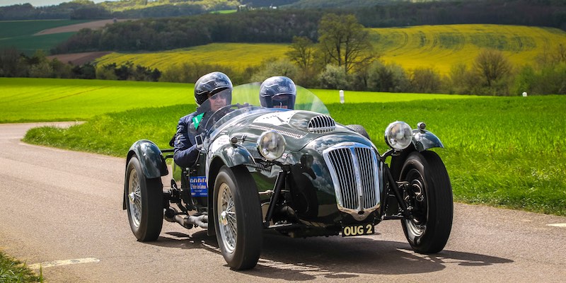 A low-riding vintage readster with two helmeted particiapnts races in the Tour Auto